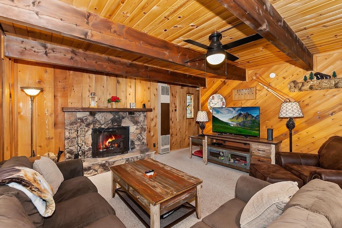 A cozy living room with a stone fireplace, wooden ceiling beams, and a television on a wooden stand.