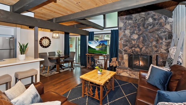 A spacious living room with a stone fireplace, wooden ceiling beams, a dining area, and a kitchen with a refrigerator.