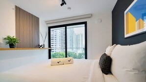 A modern hotel room with a large bed, a view of tall buildings, a wooden panel wall, and a potted plant.