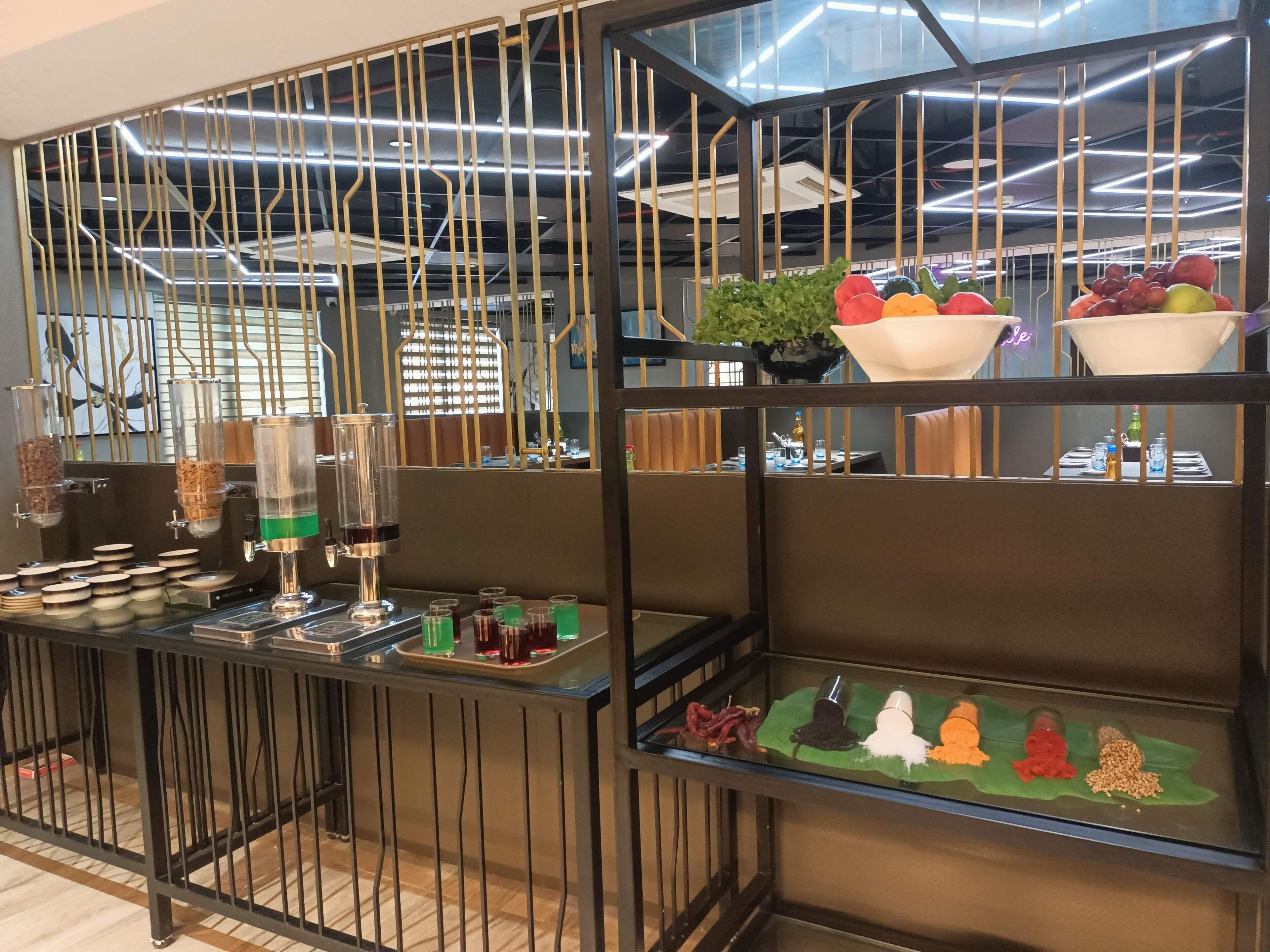 A modern bar area with a glass partition, various drink dispensers, and a selection of snacks and fruits on display.