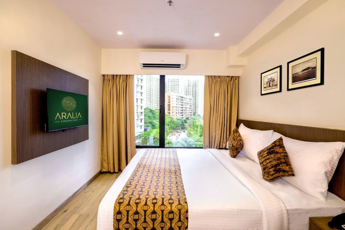 A hotel room with a bed, a mounted TV, a window with curtains, and framed pictures on the wall.