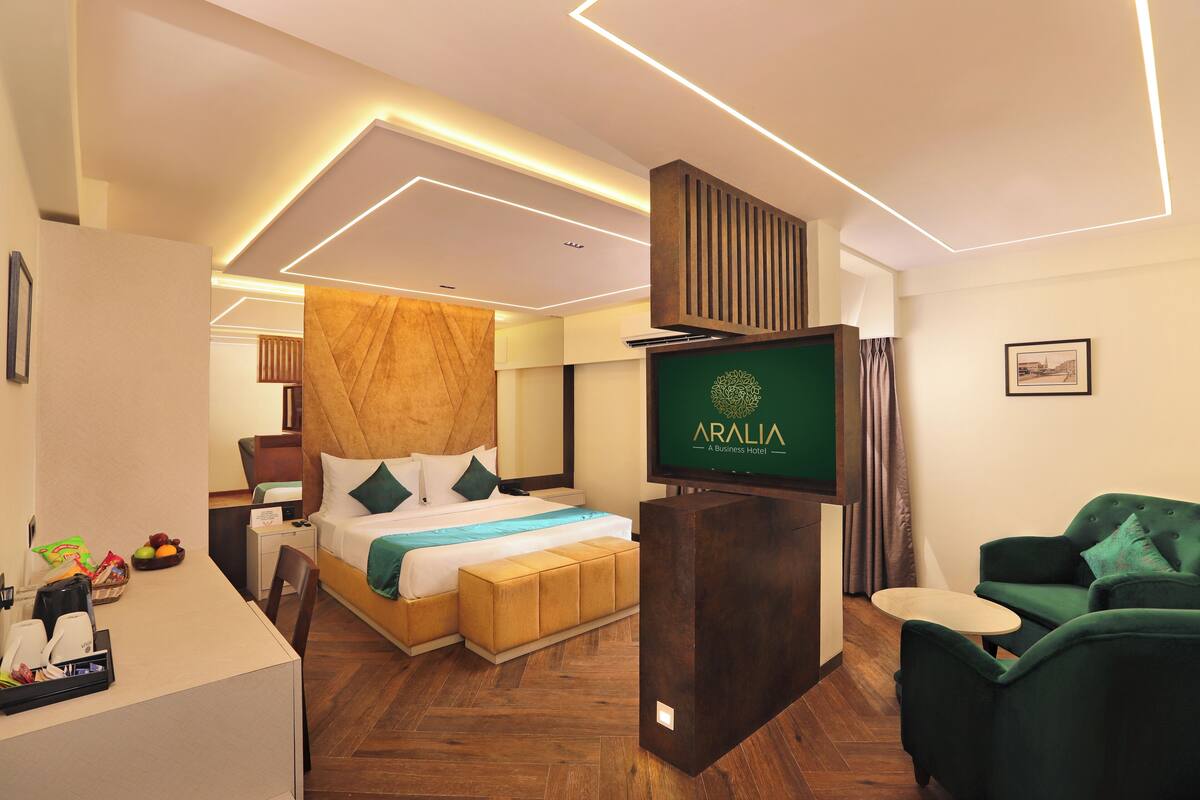 A modern hotel room with a bed, a desk, a green armchair, and a TV displaying the hotel name.