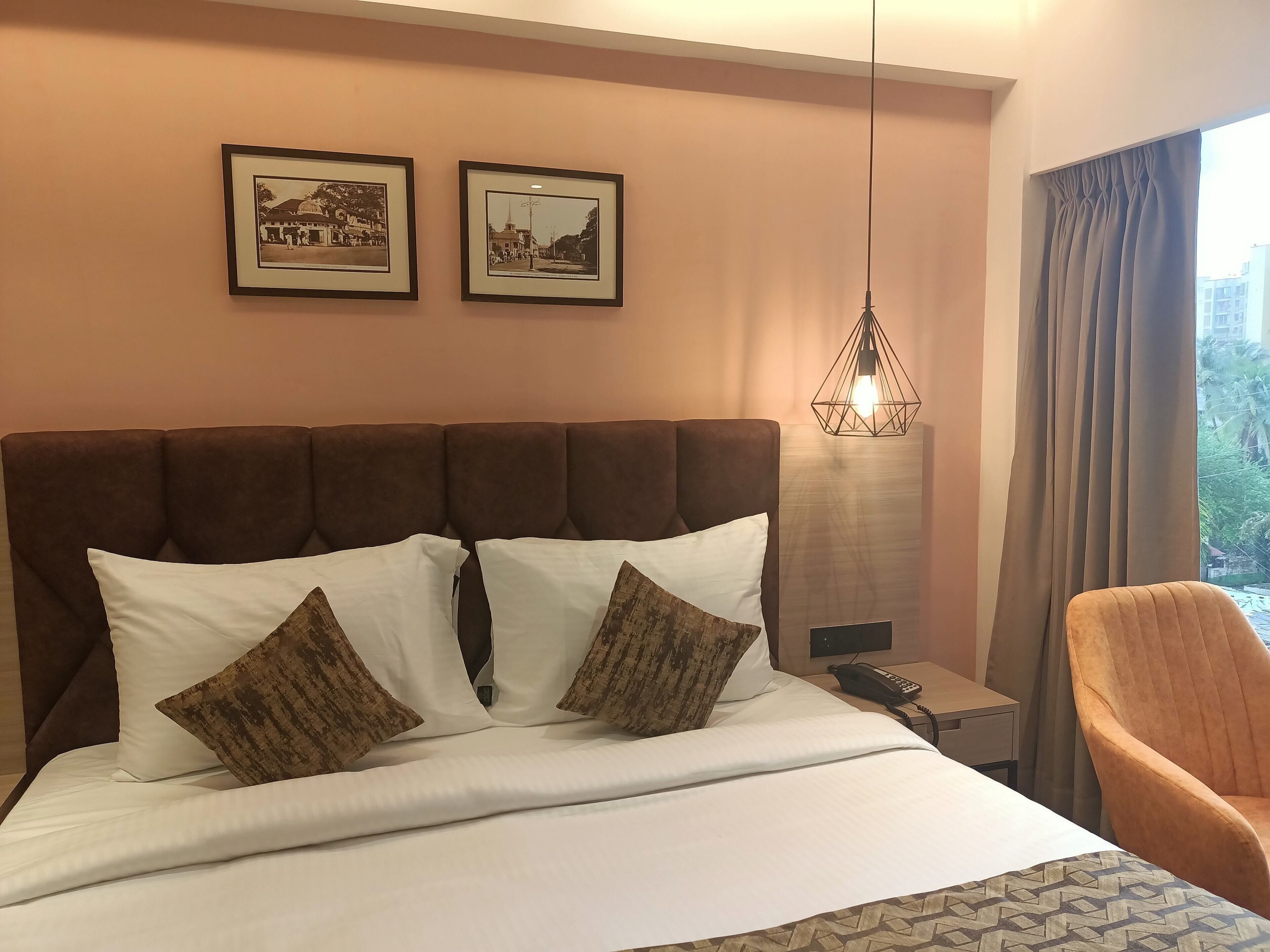 Une chambre d’hôtel avec un grand lit, deux tableaux encadrés au mur, un luminaire suspendu, une chaise et une vue sur l ’extérieur par une fenêtre.