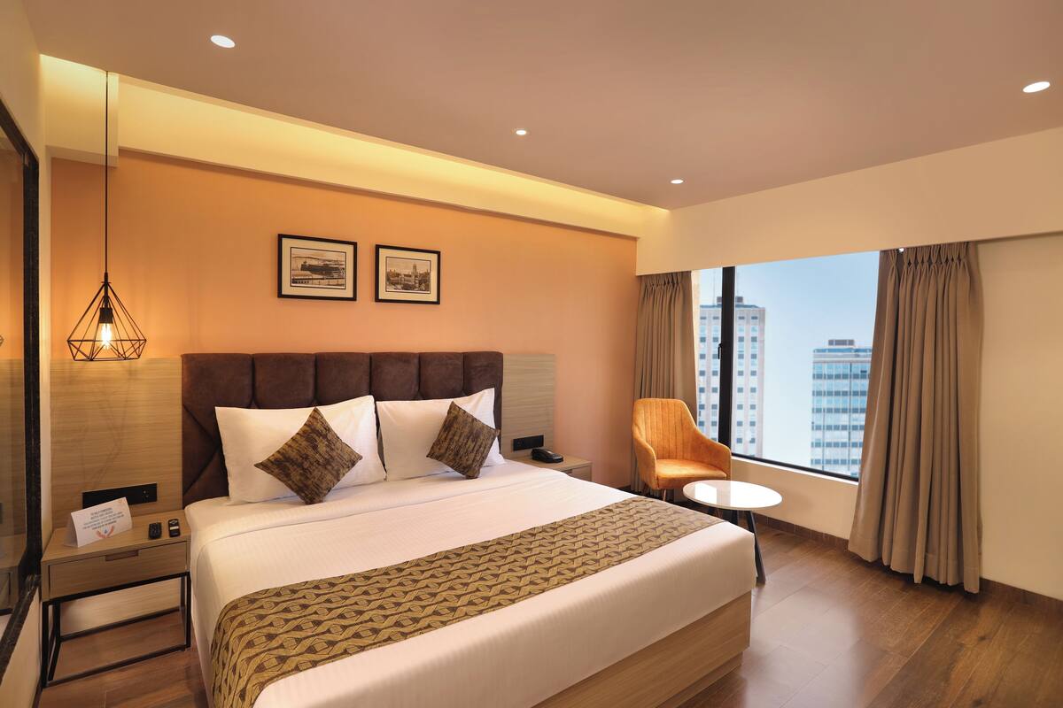 A hotel room with a large bed, a nightstand, a chair, and a window with a city view.