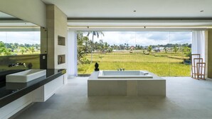 Une salle de bain moderne dotée d’une grande baignoire, d’un lavabo double et offrant une vue sur une rizière et des maisons.