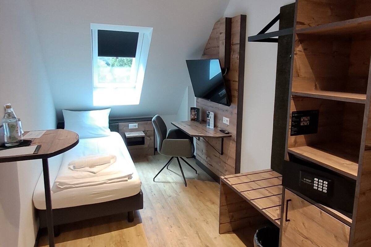 Een kleine kamer met een bed, een bureau, een televisie, een klein tafeltje met een fles en een raam.