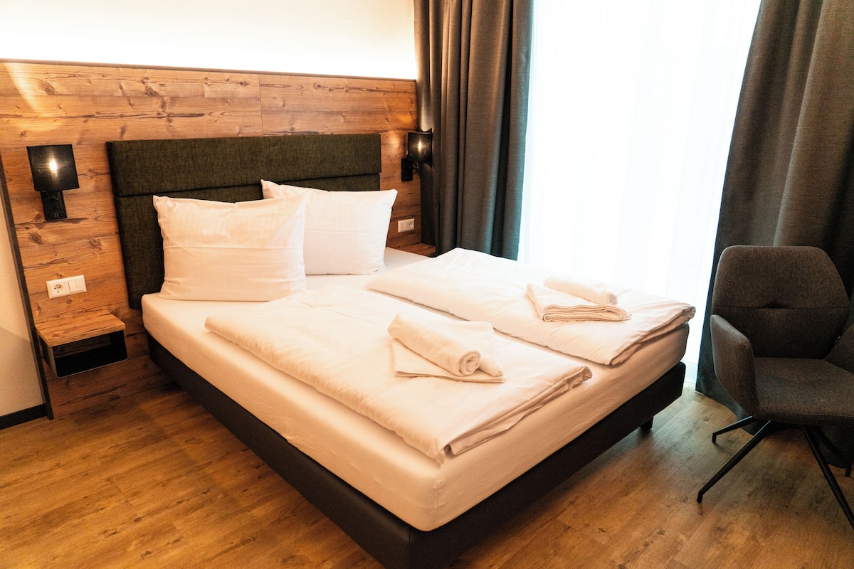 Een keurig opgemaakt bed met witte lakens en handdoeken, een houten hoofdbord en een moderne stoel.