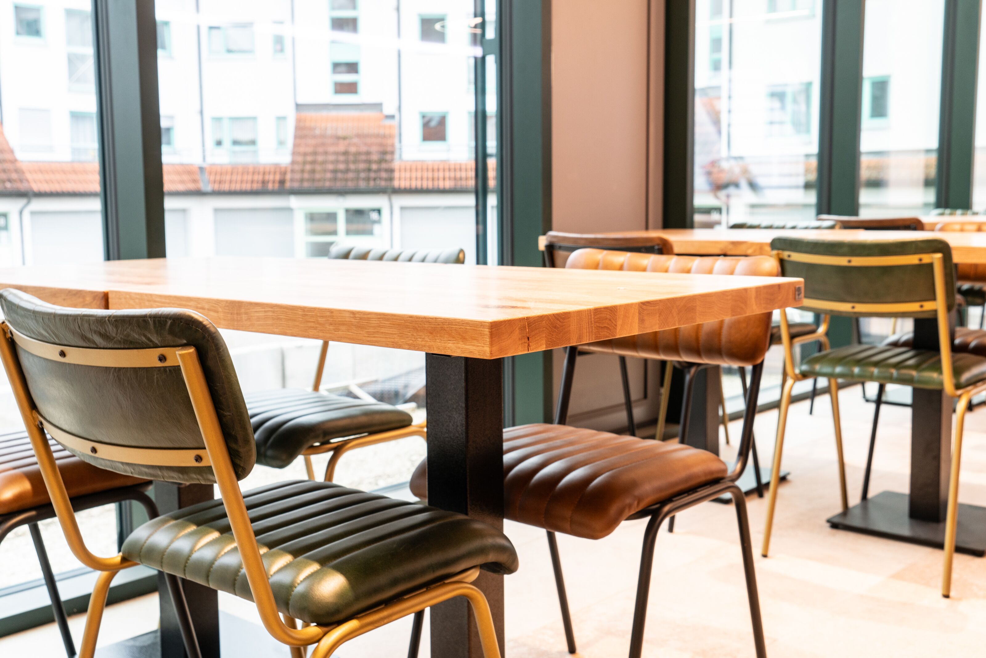 A restaurant interior with wooden tables and chairs, large windows, and a view of buildings outside.