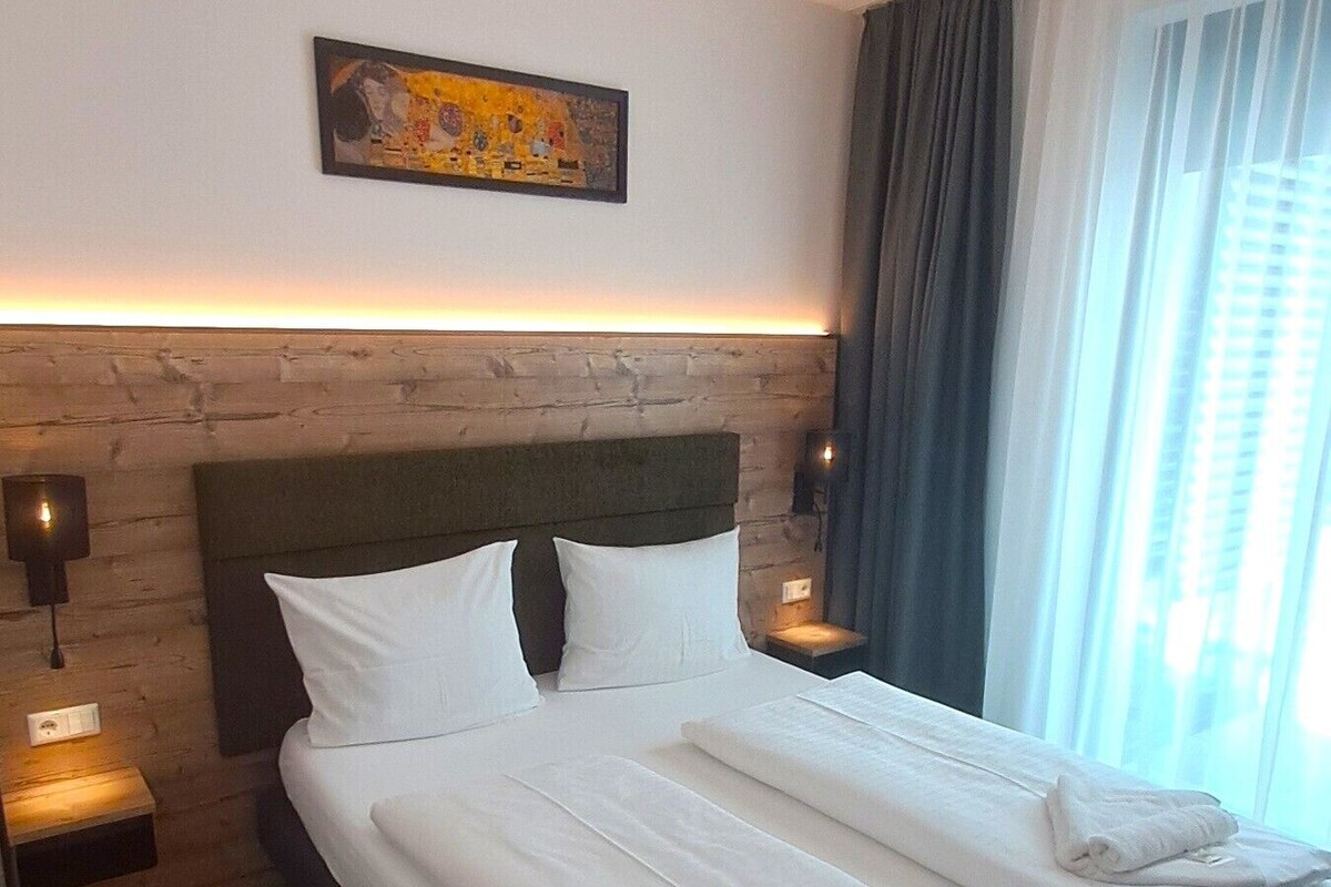 Hotelkamer met houten hoofdbord, schilderij boven het bed, twee bedlampen en een net opgemaakt bed met handdoeken.