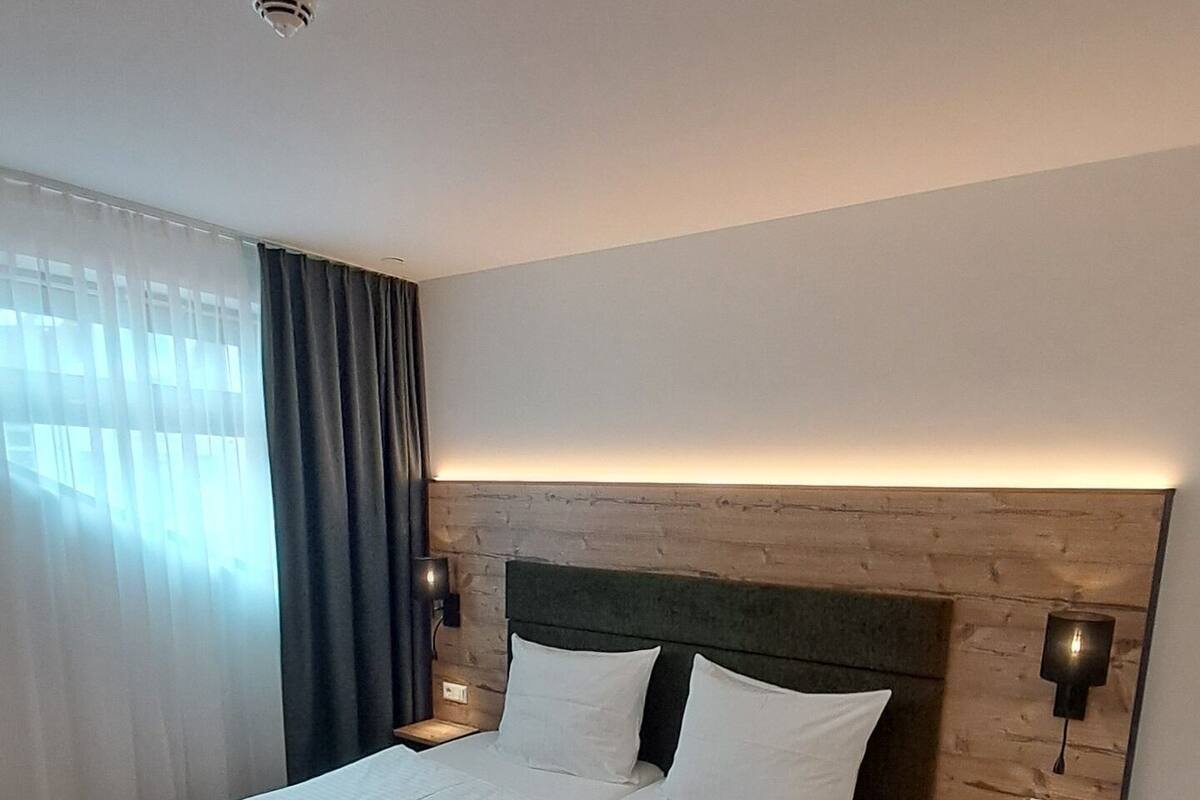 Een tweepersoonsbed met witte lakens, een houten hoofdbord, een nachtkastje met lamp en een raam met gordijnen.
