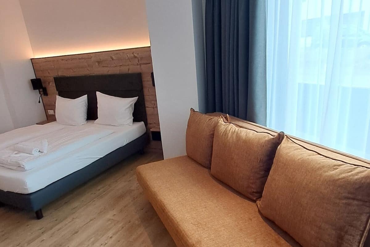 Een moderne hotelkamer met een bed, een bank, houten vloeren en een groot raam met gordijnen.