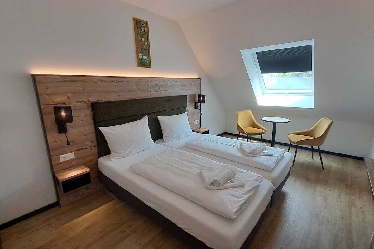 A hotel room with a wooden headboard, two beds, a small table, two chairs, a window, and a wall-mounted artwork.