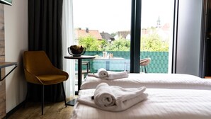 Een hotelkamer met twee bedden, een kleintje tafeltje met een schaal, een stoel en uitzicht op een groene haag en gebouwen.