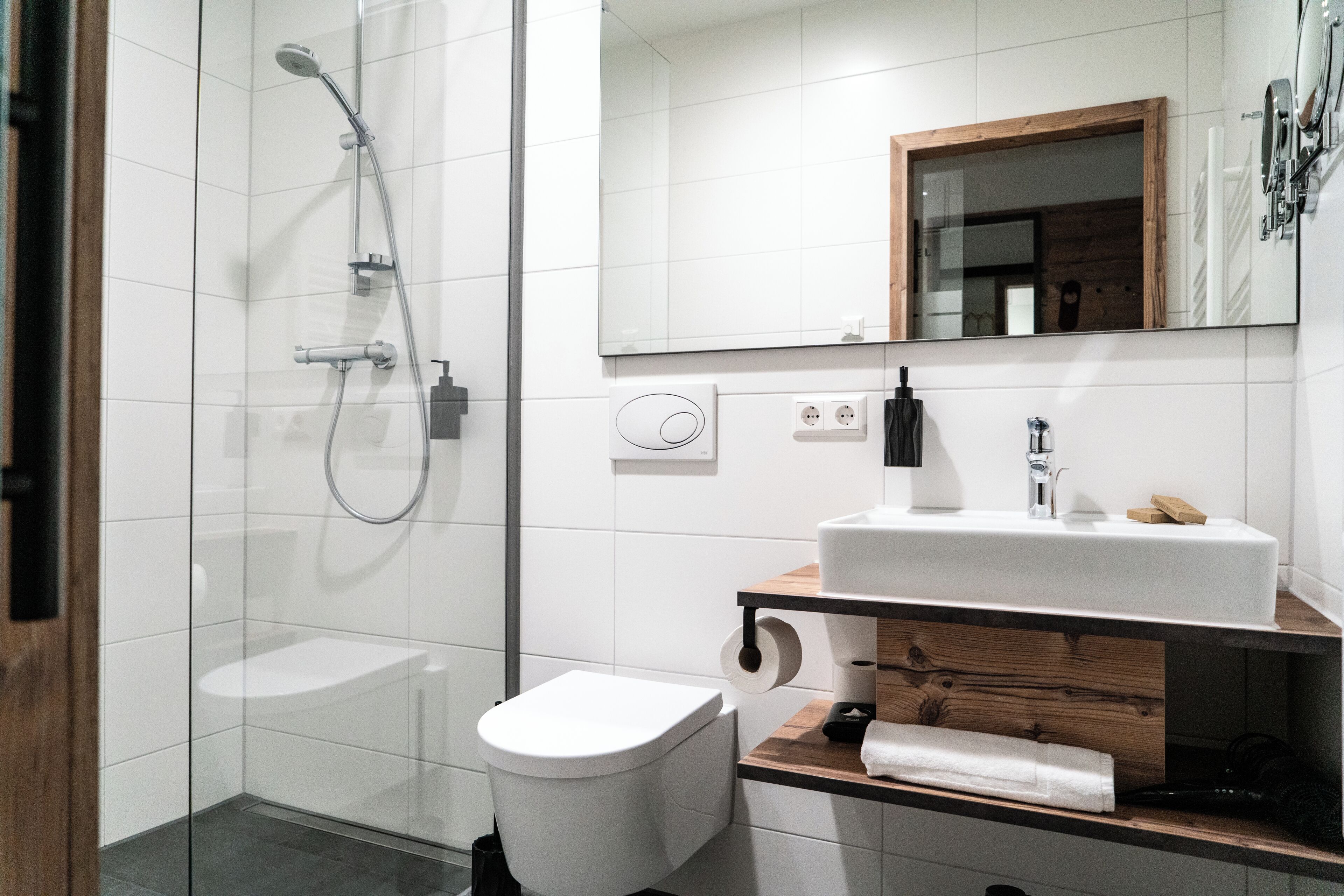 Un baño moderno con cabina de ducha de vidrio, inodoro blanco, tocador de madera con lavamanos y un espejo sobre el tocador.