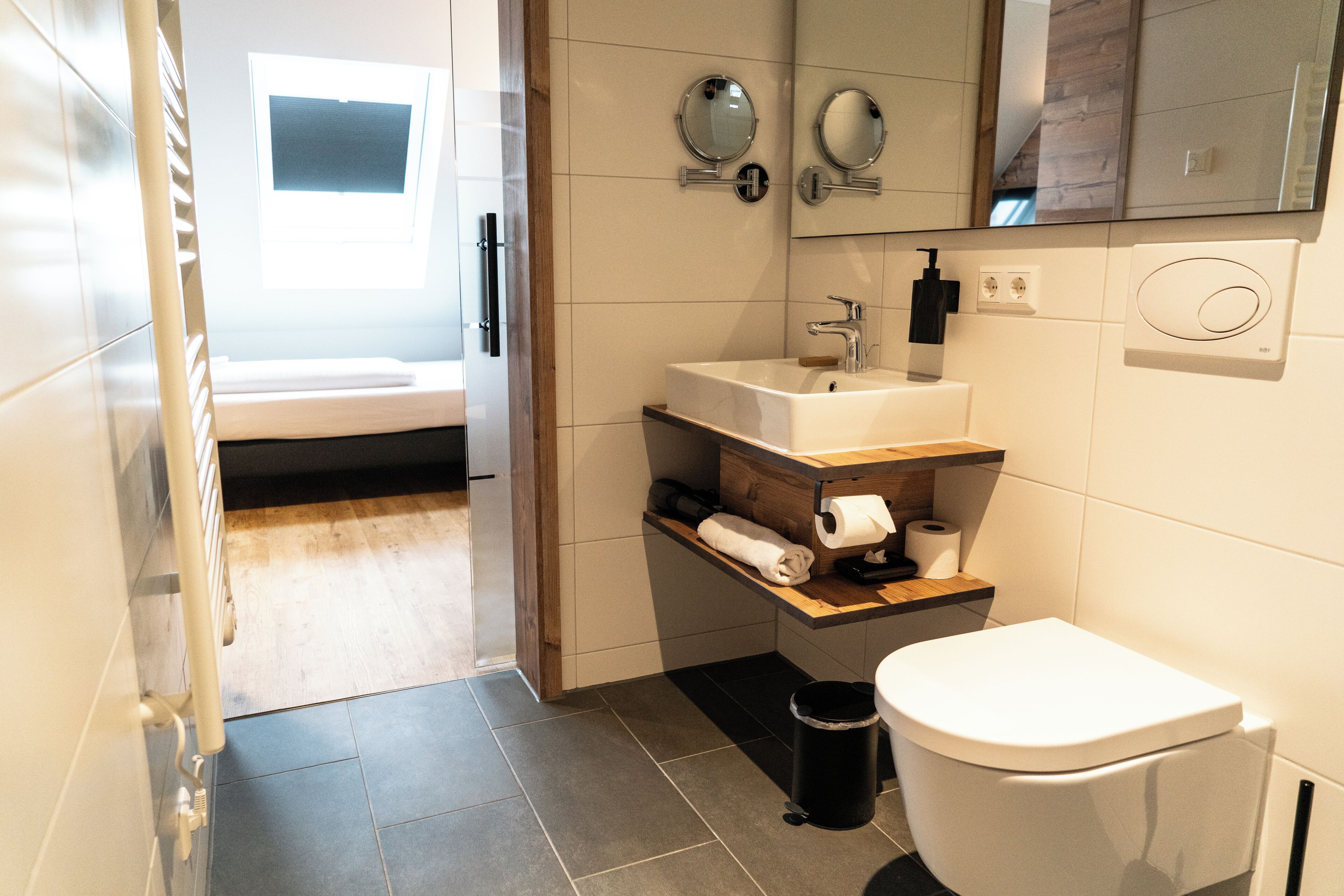 A modern bathroom with a toilet, sink, and mirror. There is a window providing natural light and a towel rack.