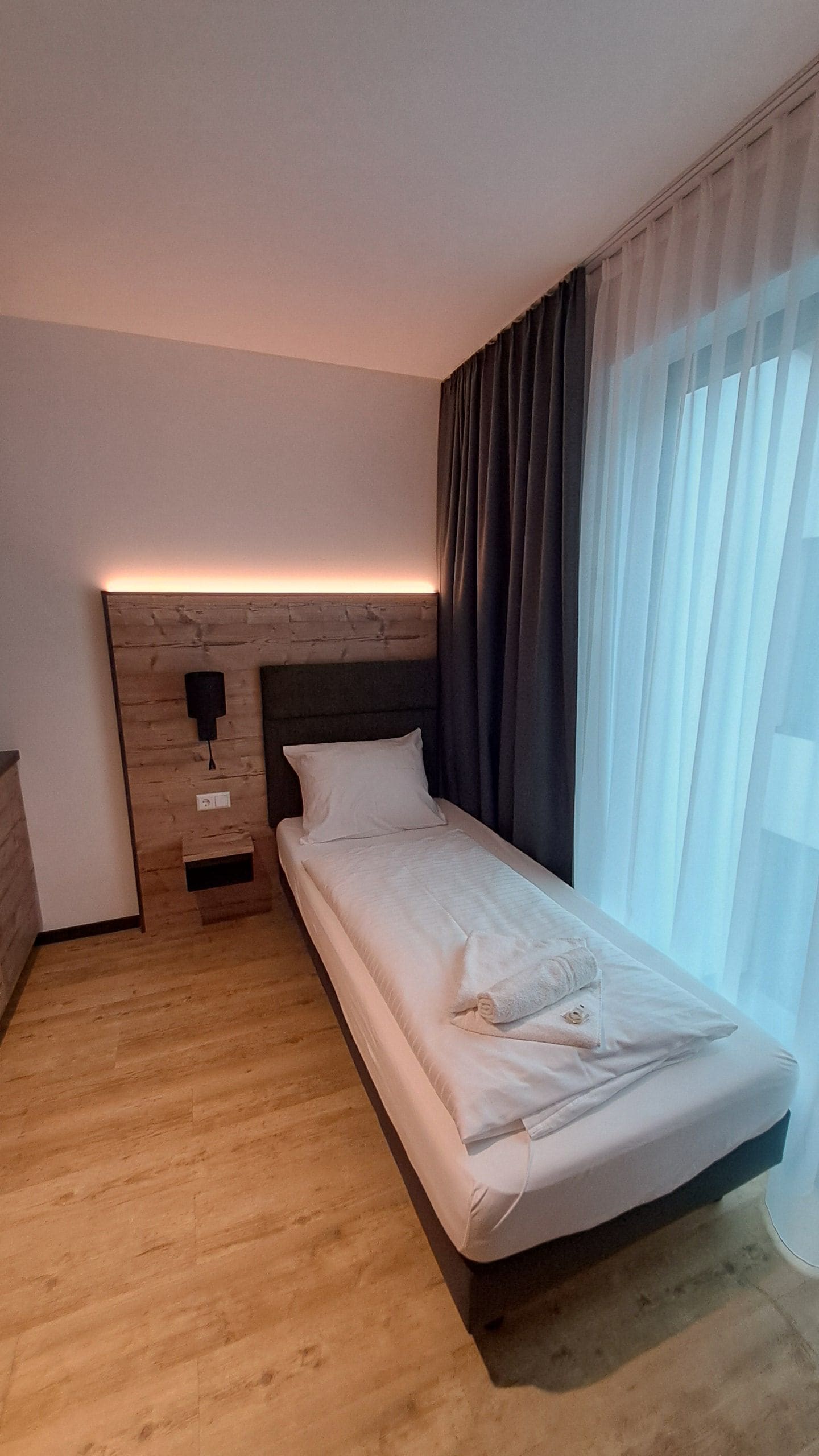 A single bed with a wooden headboard, a bedside table with a lamp, and sheer curtains on a window.