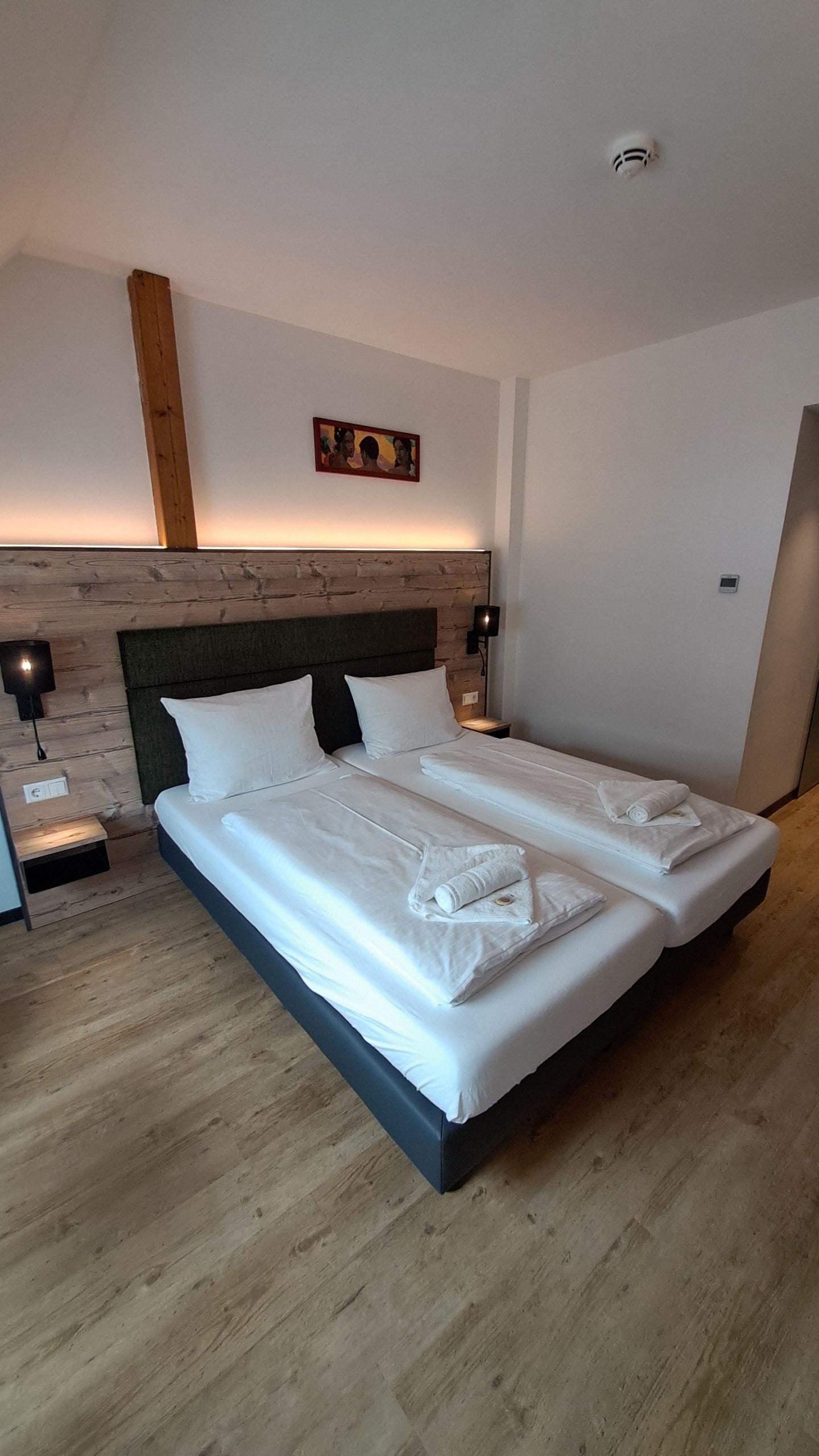 Cama doble con ropa de cama y toallas blancas, cabecera de madera, lámparas de noche y un pequeño cuadro en la pared.