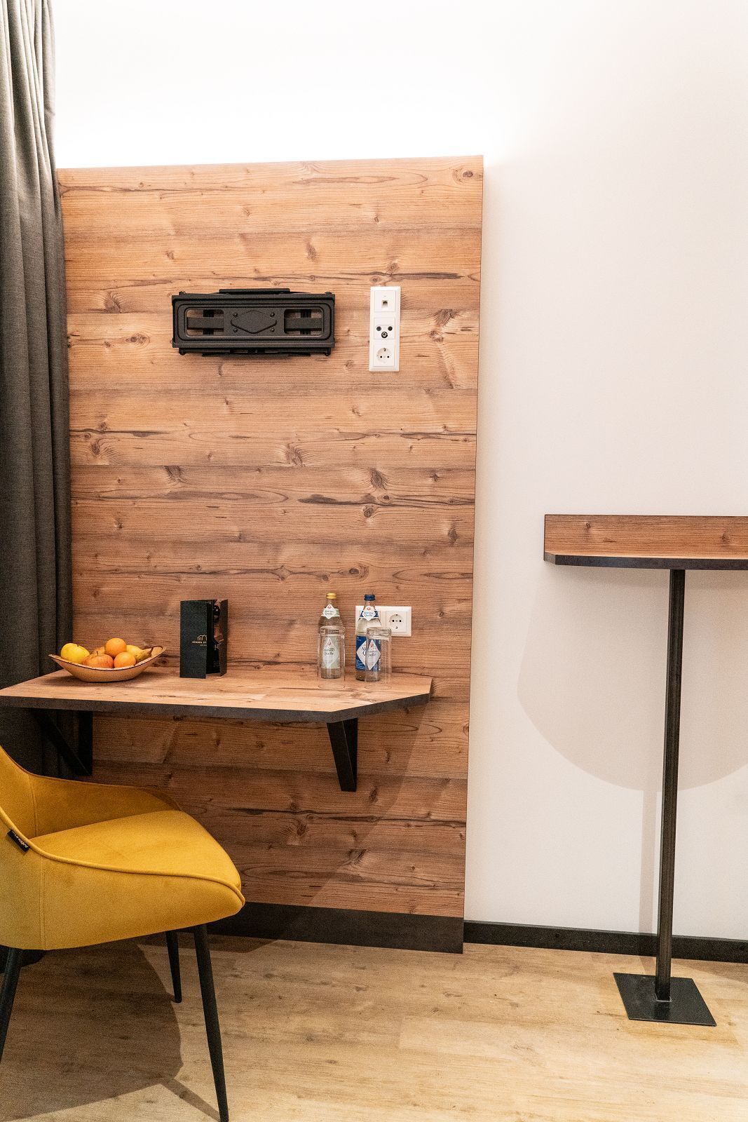 A wooden wall with a power outlet and a black electrical box. A yellow chair is positioned in front of a table with a bowl of fruit, a bottle, and a box.