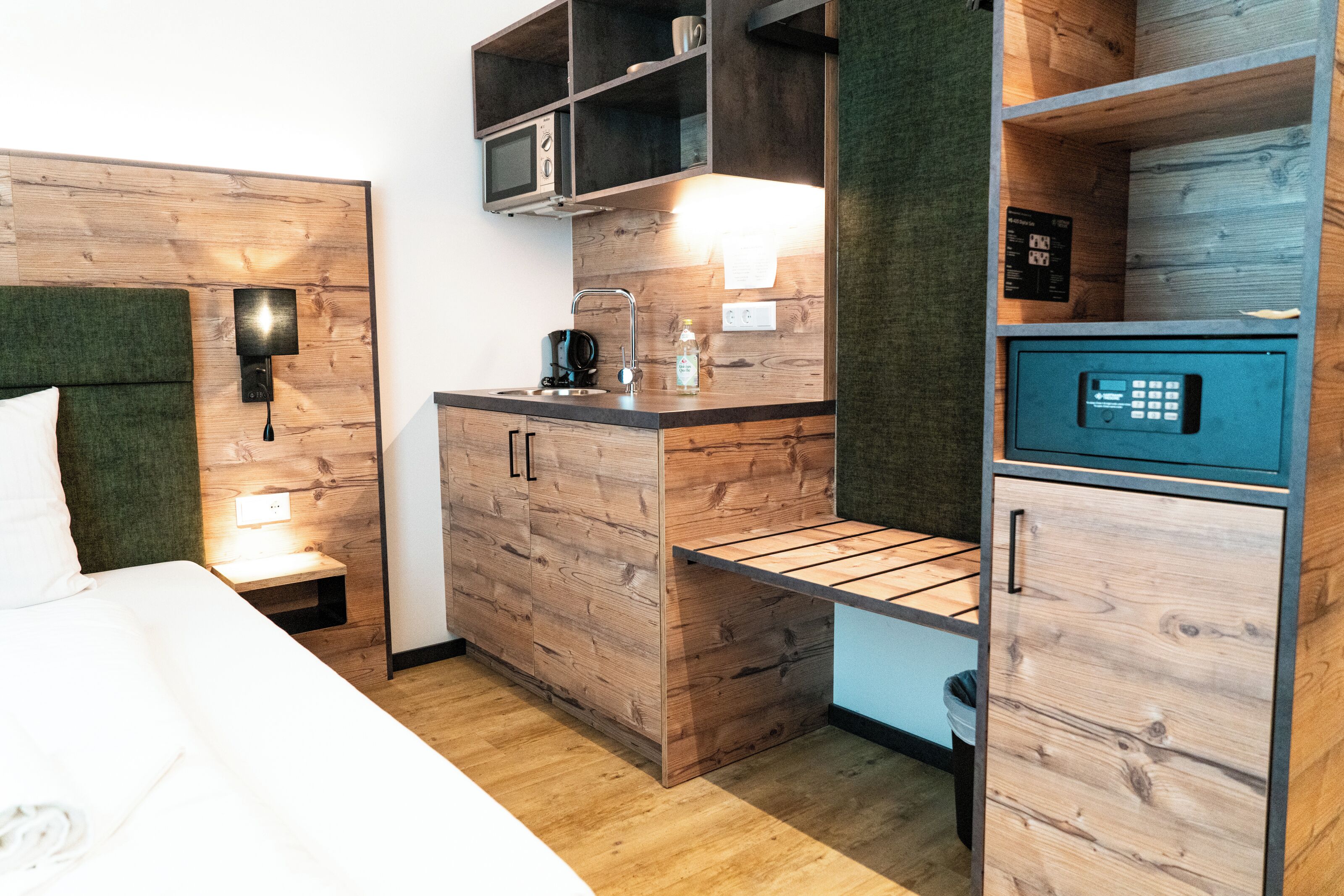 Une chambre d’hôtel compacte comprenant un lit, une kitchenette, un coffre-fort intégré et une petite étagère.