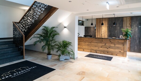 A modern hotel lobby with a wooden reception desk, a staircase with a geometric railing, and potted plants. - SEKRA Stadthotel (Heidenheim an der Brenz)