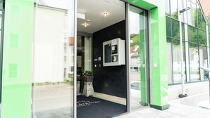 Een hotelingang met een glazen deur, een receptiebalie en een zwart tapijt met een logo.