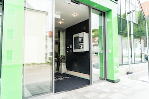 Een hotelingang met een glazen deur, een receptiebalie en een zwart tapijt met een logo.