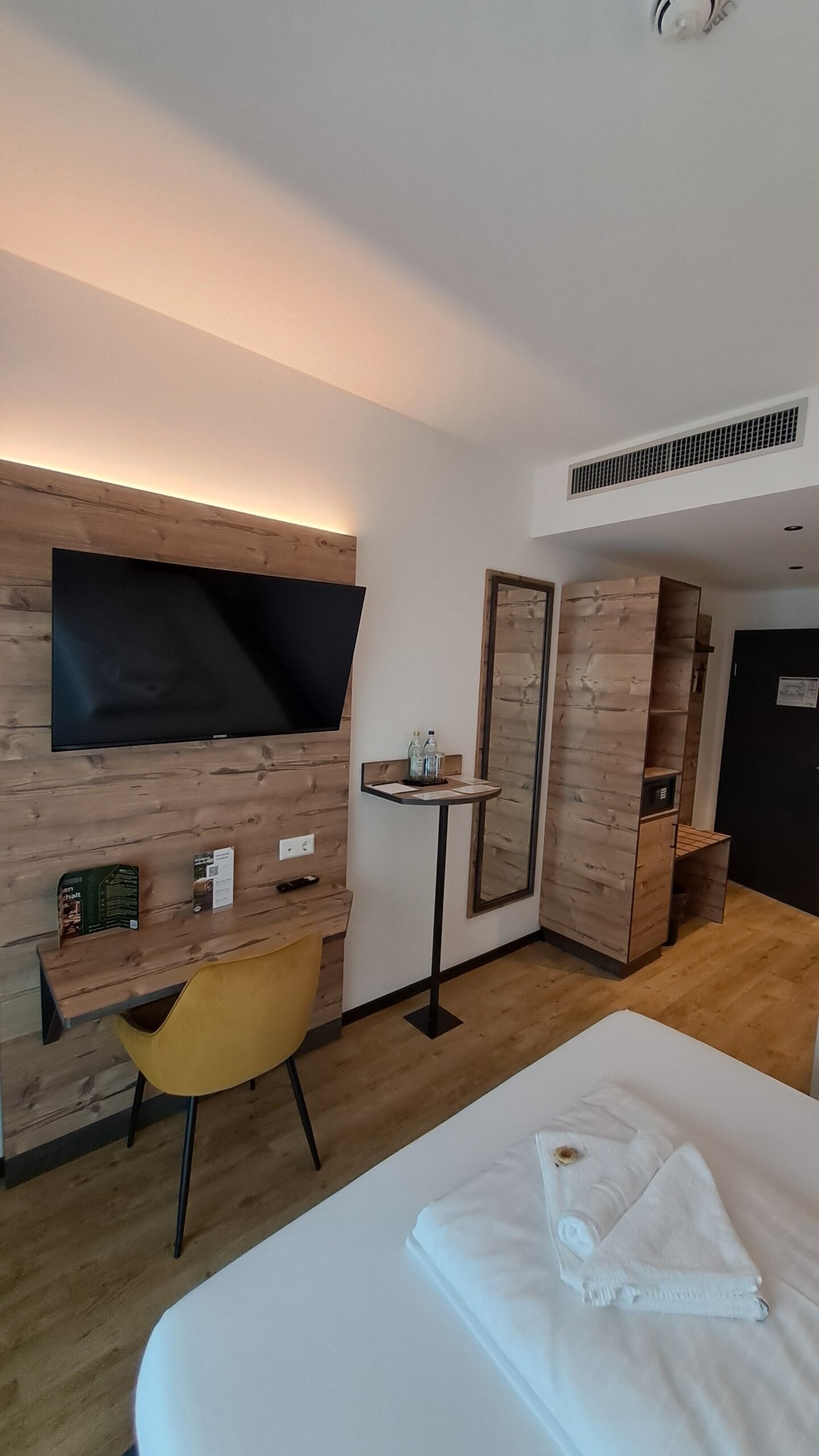 Habitació d'hotel amb un llit, una taula de treball, una cadira, un televisor, un mirall i una taula petita amb ampolles.