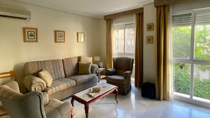 A living room with a sofa, armchair, and coffee table, a window with curtains, and framed pictures on the wall. - Rentalsevilla en Nervión (Sevilla)
