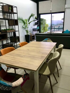 A modern interior with a wooden table, chairs, a bookshelf, a potted plant, and a view through a large window. - Luxury 1br-2br Apt-panoramic View Terrace 24-7 Pwr (Ain Saadeh)