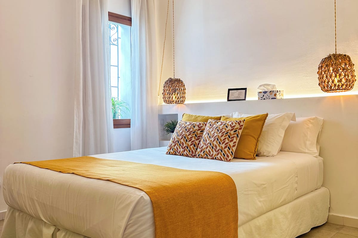 Un dormitorio con una cama grande, vigas de madera en el techo, una ventana con cortinas y dos lámparas colgantes.