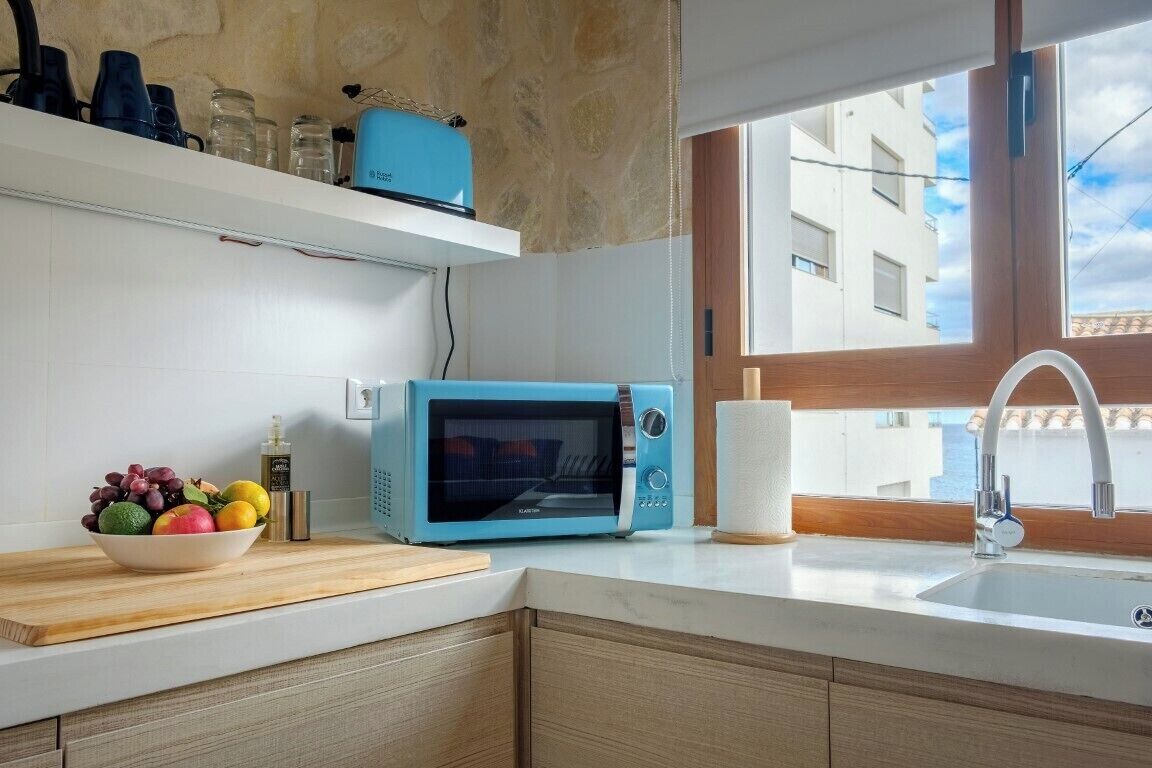 A kitchen with a blue microwave, a bowl of fruit, and a window with a view of buildings.