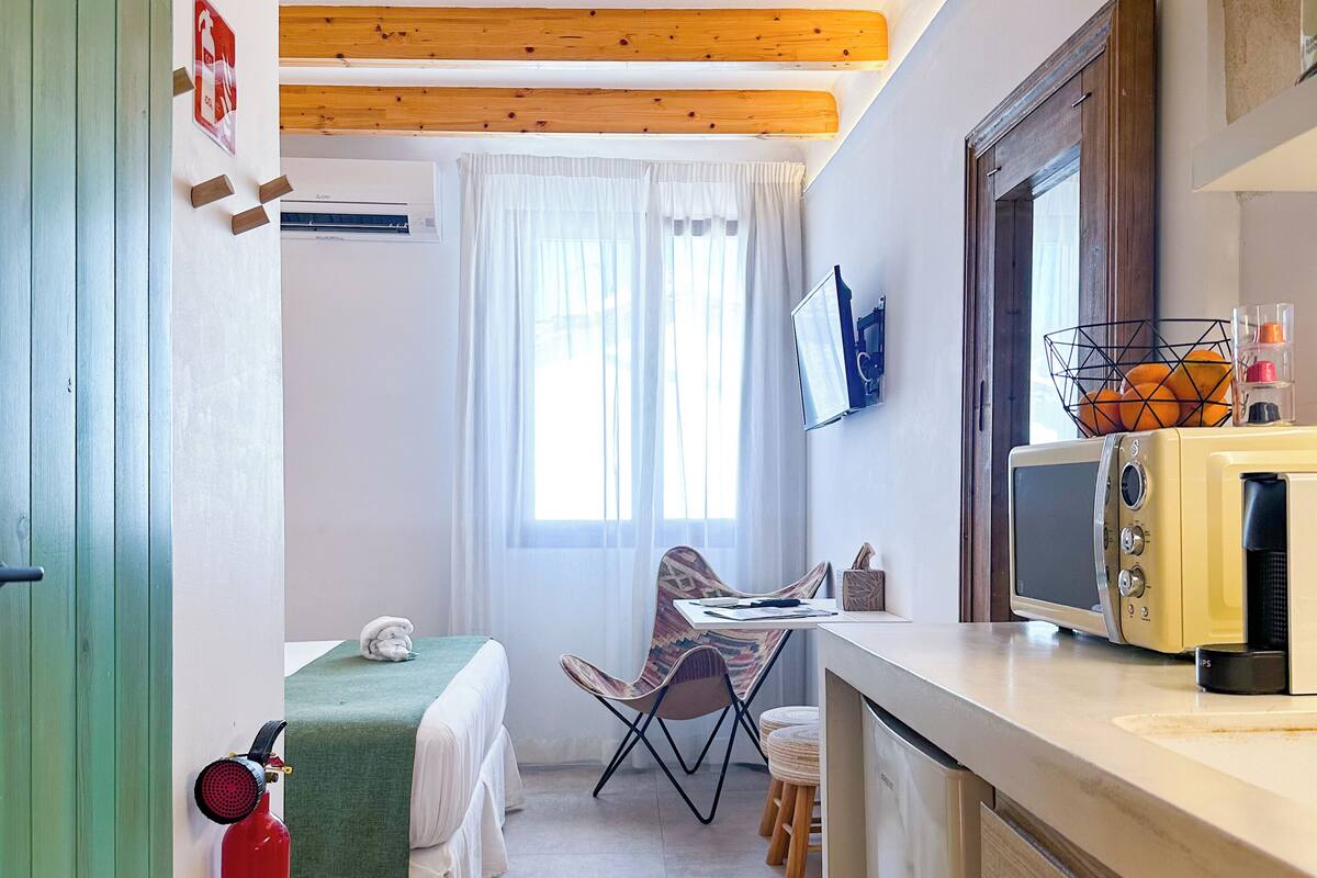 Una habitación compacta con cama, una mesita, una silla, microondas y extintor.