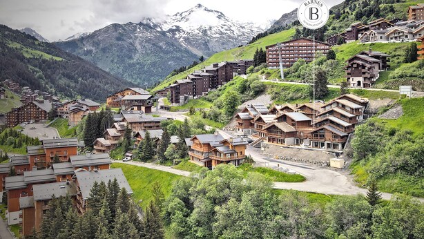 A mountainous resort with wooden buildings, a ski lift, and snow-capped peaks in the background.