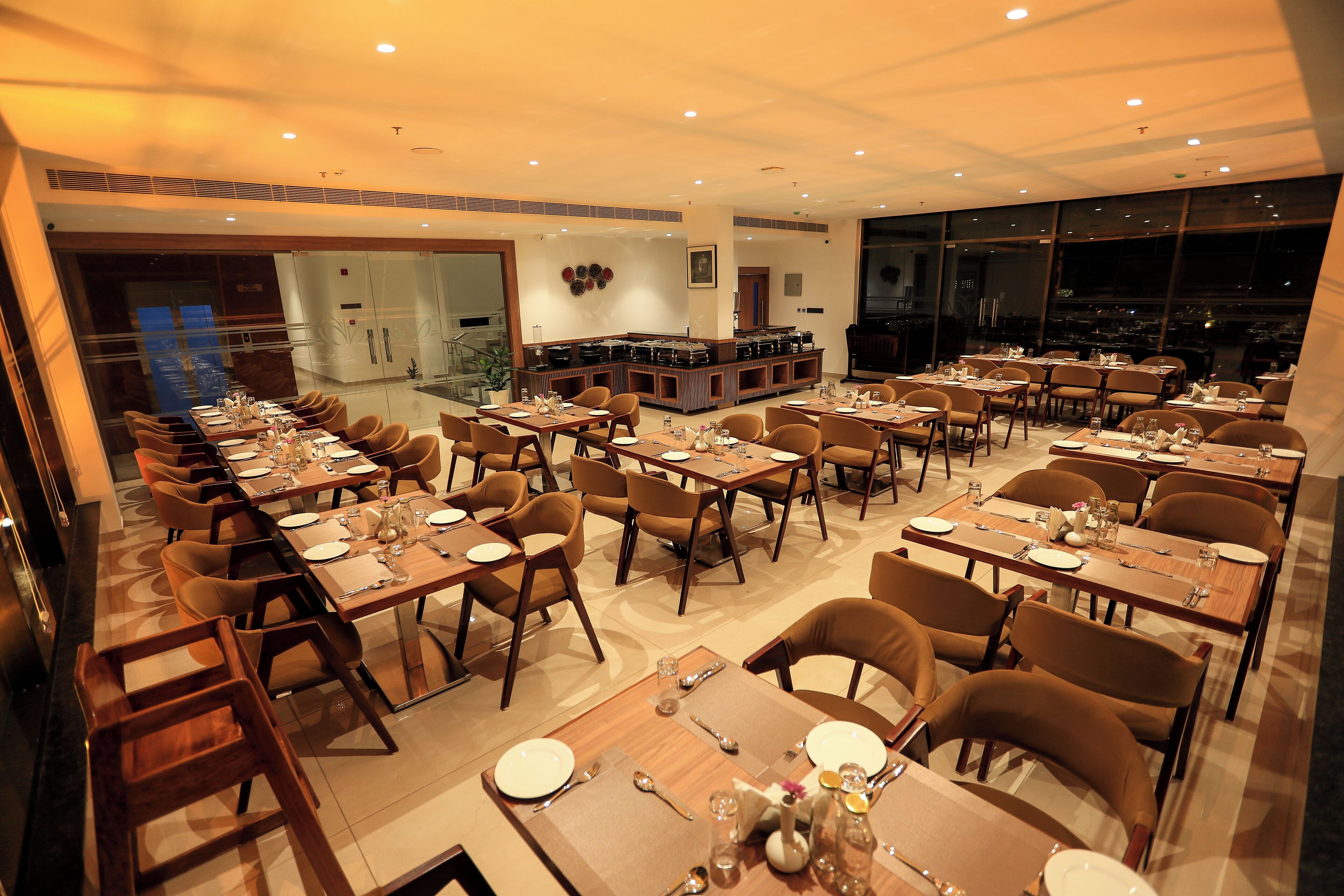 A spacious dining area with multiple tables and chairs, a central bar, and large windows offering a city view.