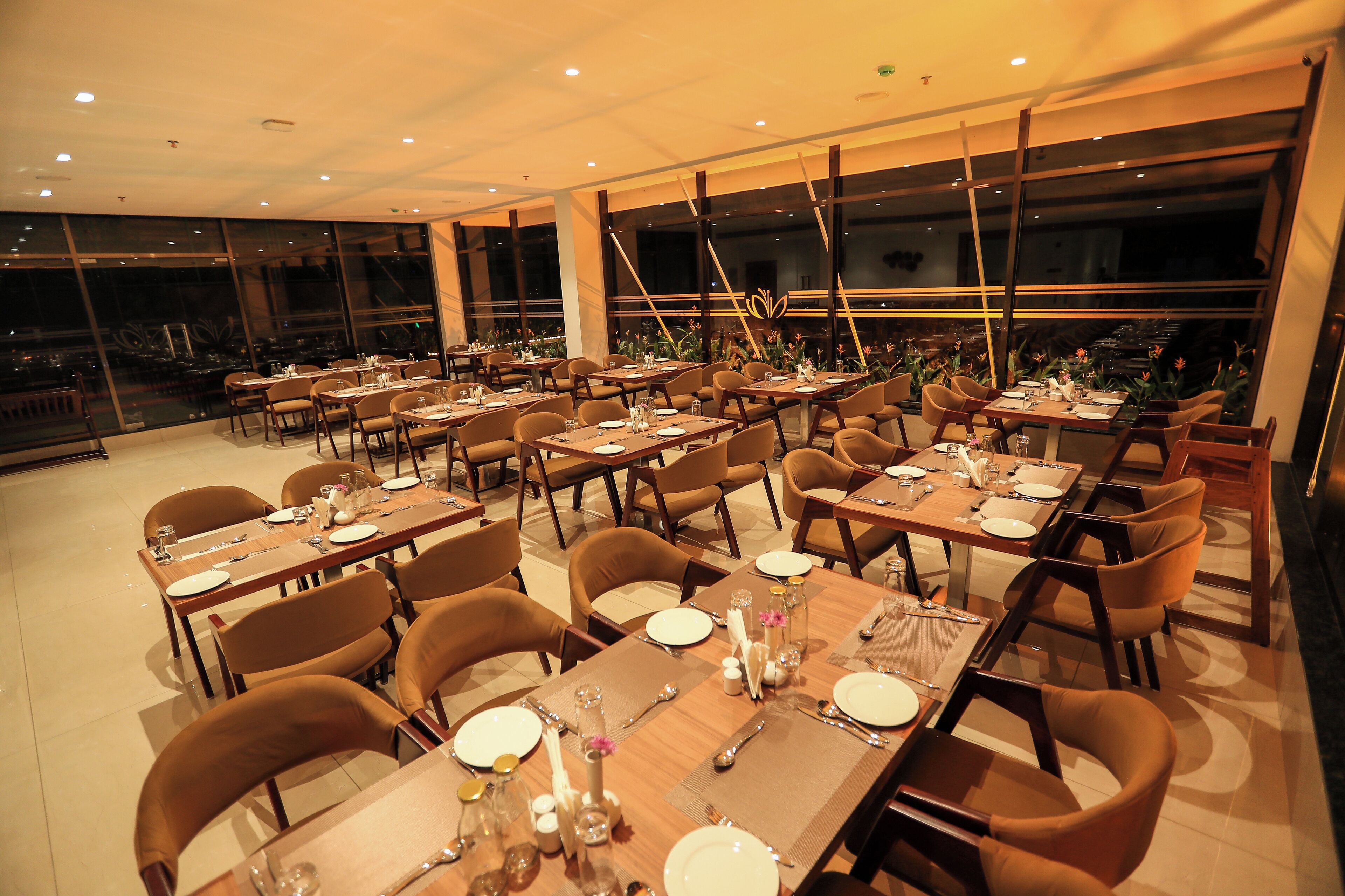 Um restaurante com várias mesas e cadeiras, dispostas em um espaço amplo com grandes janelas que oferecem vista para a paisagem urbana.