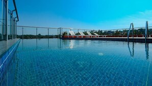 A clear blue swimming pool with tiled flooring, a metal railing, and a row of sun loungers with red cushions. - Hotel Saffron Wayanad (Sulthan Bathery)