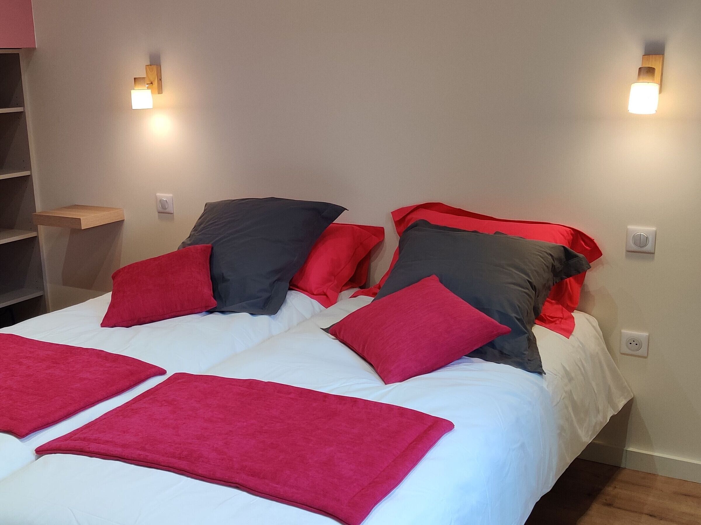 A double bed with red and grey pillows, a wooden shelf, and two wall-mounted lights.