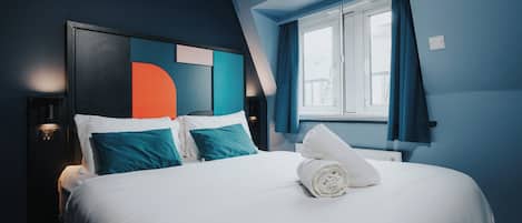A neatly made bed with white linens and a rolled towel, a modern headboard with a geometric design, and a window with blue curtains.