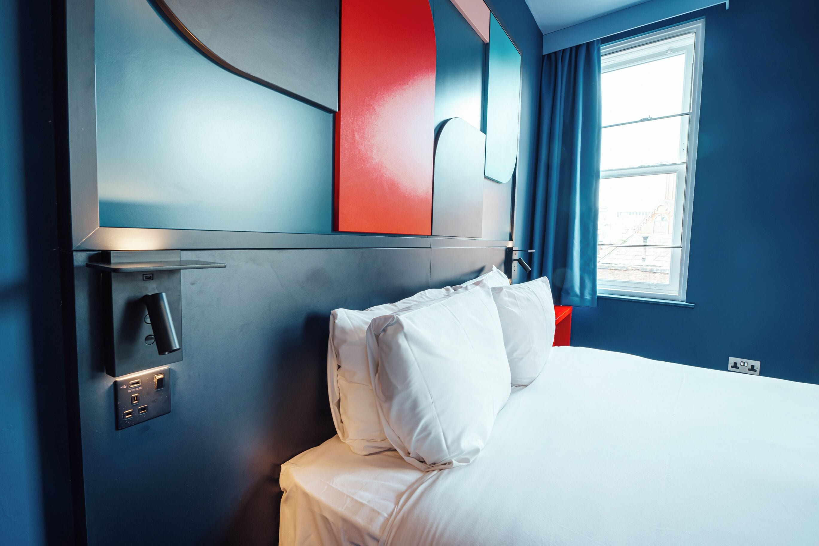 A hotel room with a bed, a bedside table, a power outlet, and a window with curtains.