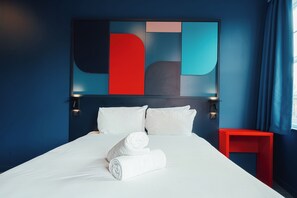 Quarto de hotel com uma cama grande, almofadas brancas, uma mesinha de cabeceira vermelha e uma obra de arte abstrata moderna na parede.