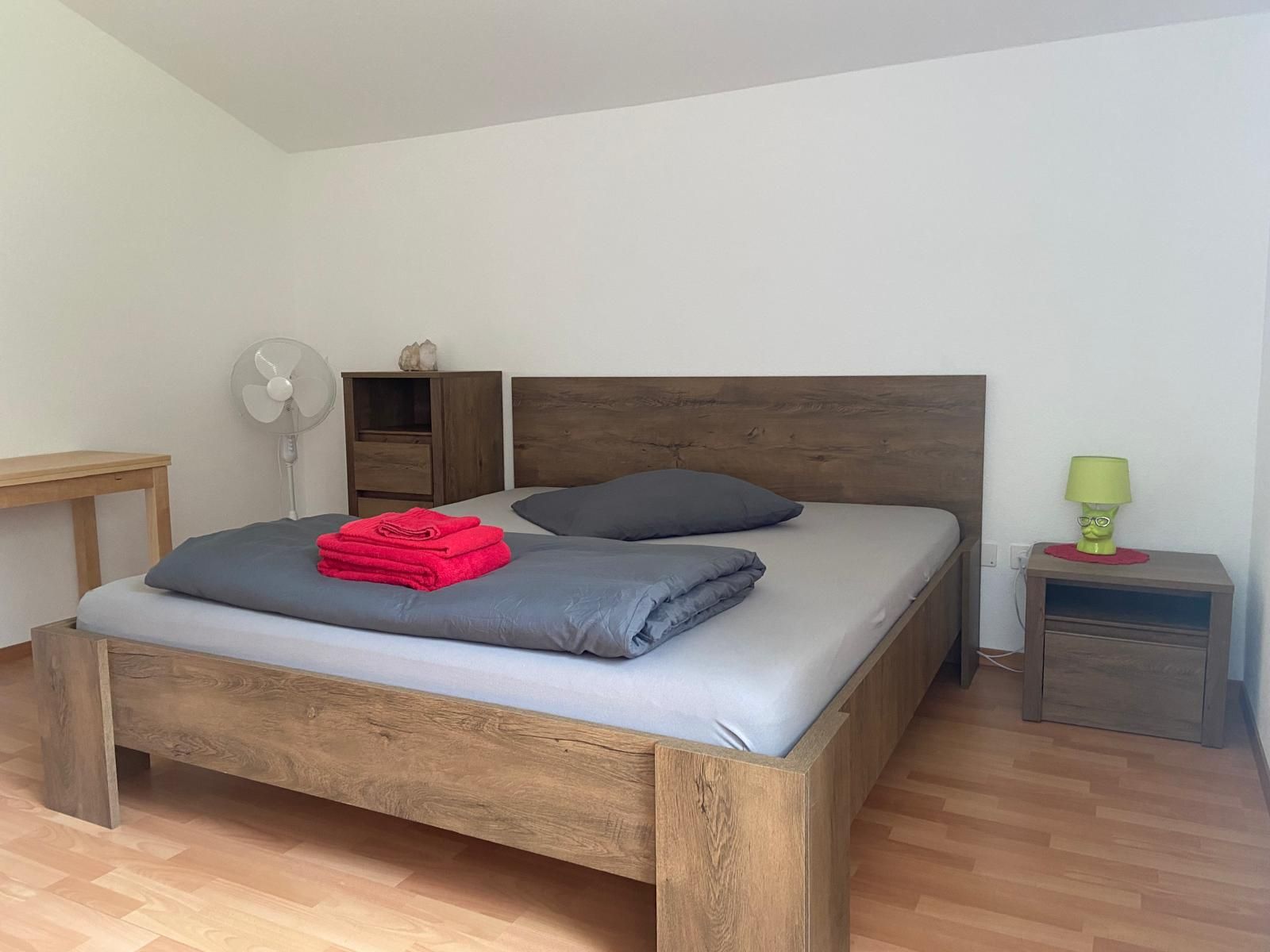 Une chambre avec un lit simple, une tête de lit en bois, une table de chevet avec une lampe, une petite armoire et un ventilateur.