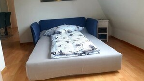 Une chambre avec un lit simple, un plafond en pente, un tableau représentant un paysage, une table de chevet et une petite table de nuit.