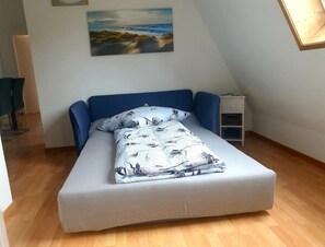 Une chambre avec un lit simple, un plafond en pente, un tableau représentant un paysage, une table de chevet et une petite table de nuit.
