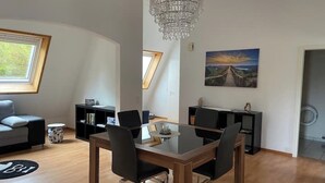 Ein modernes Wohnzimmer mit Esstisch, Stühlen, einer Deckenleuchte, einer Couch, einem Bücherregal und einem Gemälde.