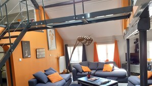 A modern living room with a staircase, grey sofas with orange cushions, a coffee table, and a large floor lamp. - Le Toit de Charme D'isabelle ! (Beaune)