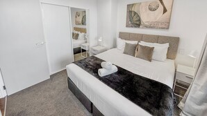 A modern hotel room with a large bed, a bedside table with a lamp, a mirror, and a painting on the wall. - Ellerslie Sanctuary | Luxury Apt & Great Amenities (Auckland)