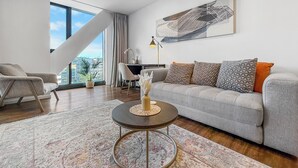 A modern living room with a grey sofa, a round coffee table, and a large window offering a city view. - Ellerslie Sanctuary | Luxury Apt & Great Amenities (Auckland)