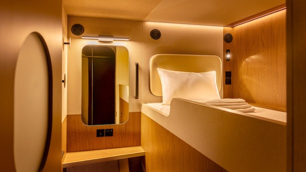 A compact, well-lit bathroom with a wooden vanity, a mirror, and a bed with a white pillow. - Kabannas London St Pancras (London)