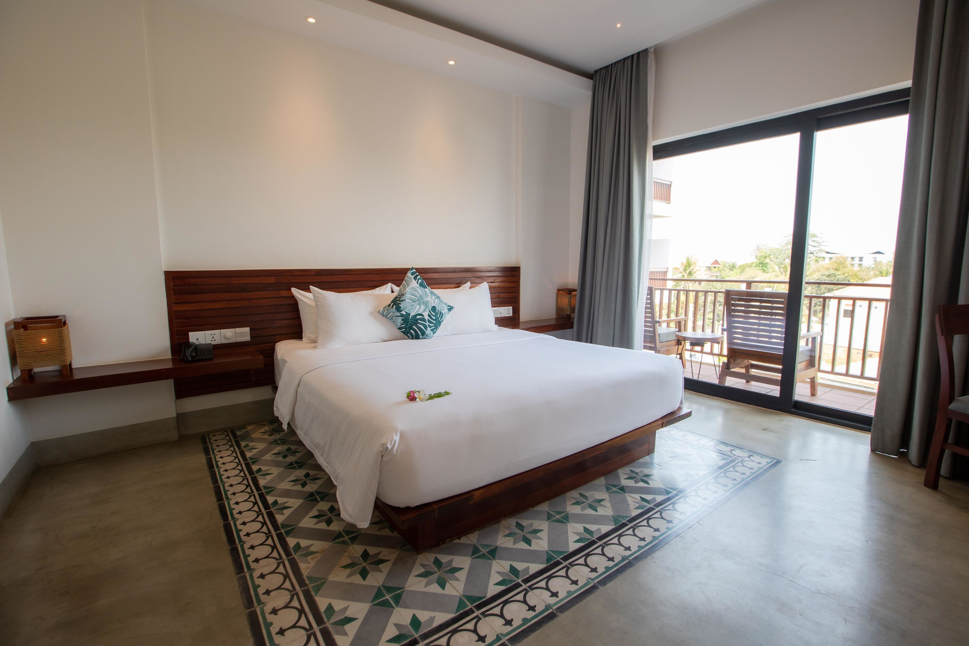 A hotel room with a large bed, a wooden headboard, a patterned rug, and a balcony with a view.