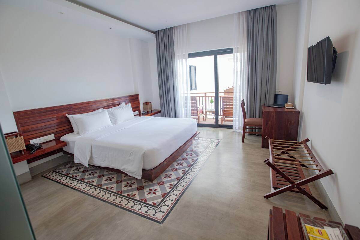 A hotel room with a large bed, a desk with a laptop, a TV mounted on the wall, and a balcony with a view.